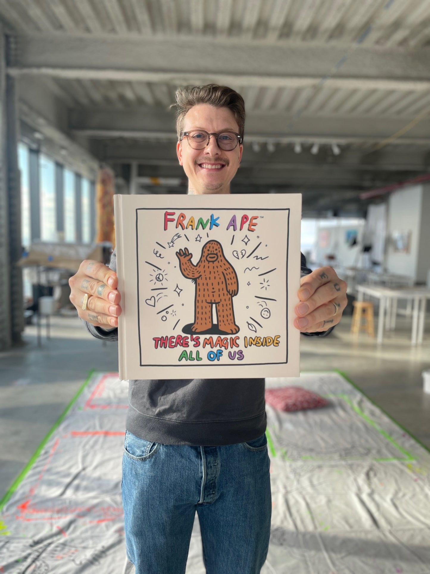 Artist Brandon Sines holding his hardcover book titled 'There's Magic Inside All of Us' with a colorful illustration of Frank Ape, a magical sasquatch on the cover.