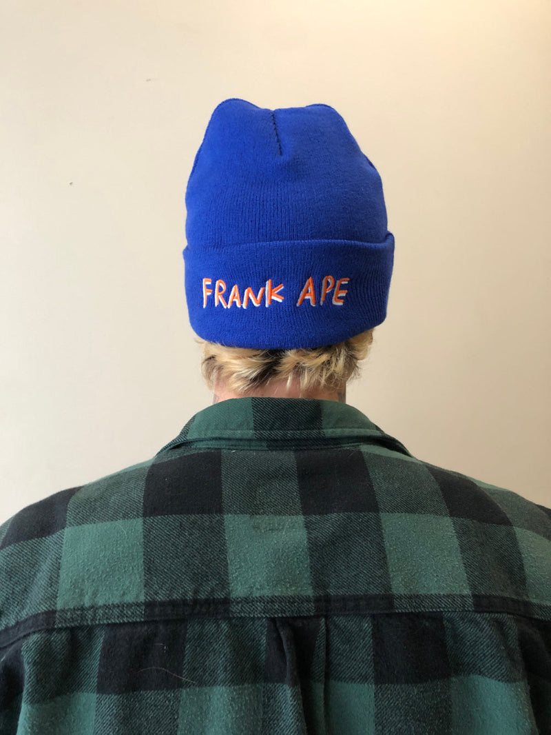 Artist Brandon Sines wearing a Frank Ape beanie. Frank Ape double sided embroidered beanie. This photo shows the side with Frank Ape signature text. Blue hat with white and orange stitching on this side.